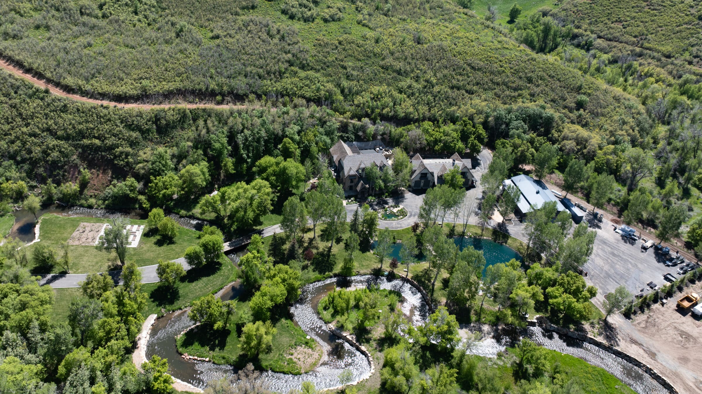 Aerial view of a large estate ideal as a hunting property or cattle ranch, featuring a stone house, landscaped lawns, winding creek, pond, trees, parking area, and open green fields surrounded by dense forest.