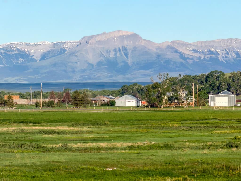 Green fields and a rural area with houses and farm buildings at Baker Spring Creek Ranch, set before large, snow-dusted mountains under a clear blue sky.