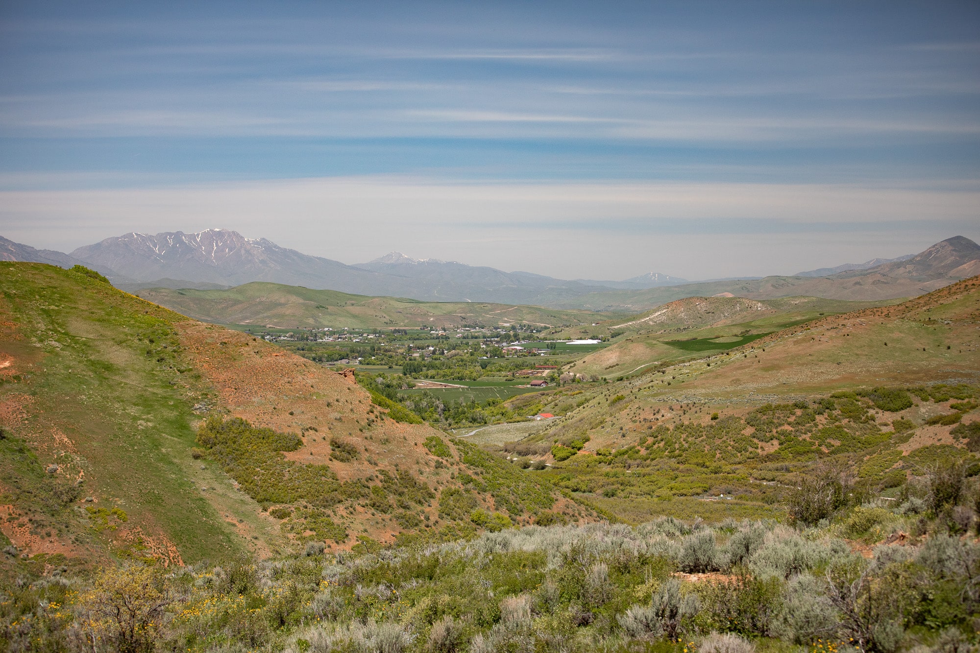 A wide, green valley stretches between rolling hills under a blue sky with wispy clouds, distant mountains rising in the background and a small town nestled amid trees—an ideal recreational land or hunting property.
