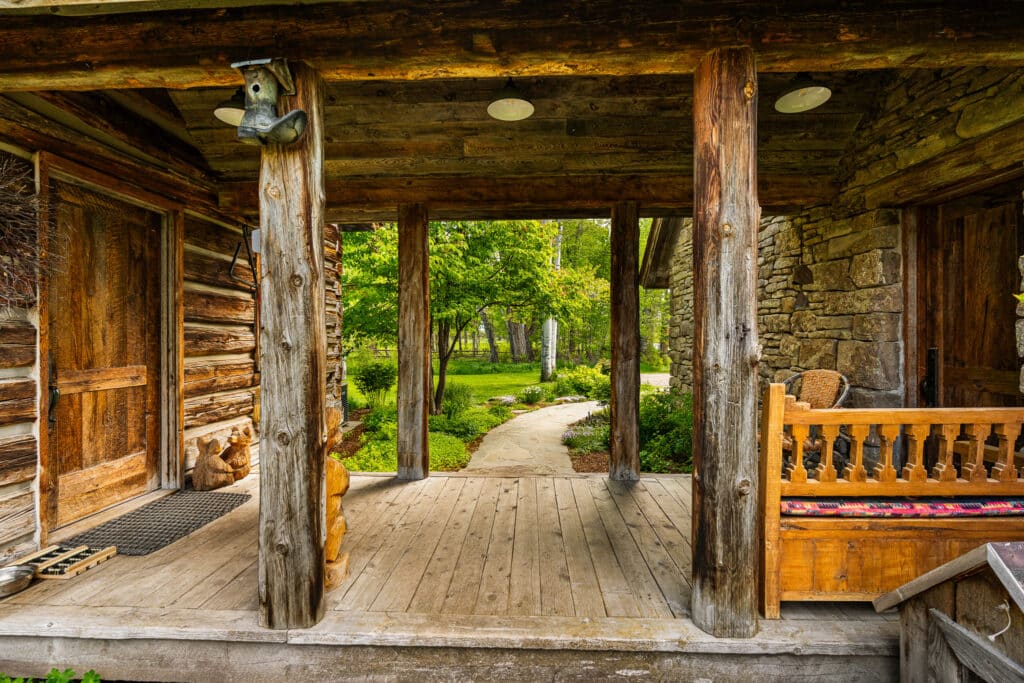 A rustic wooden porch with log pillars and a bench overlooks a garden path leading to green trees and grass—perfect for enjoying nature on this beautiful land for sale.
