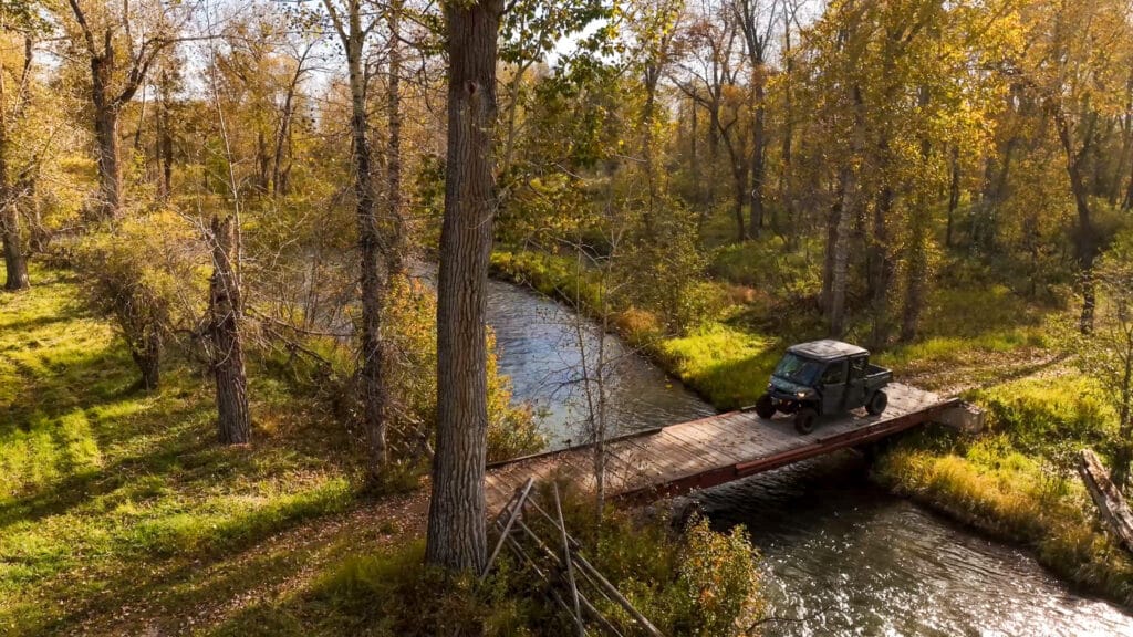 A black ATV is parked on a small wooden bridge over a winding creek in a forest with tall autumn trees, the sunlight casting a warm glow—an inviting scene for those seeking recreational land or exploring ranch for sale opportunities.