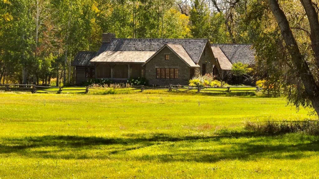 A stone cottage with a dark roof sits surrounded by trees and greenery, bordered by a wooden fence and a wide, sunlit grassy lawn—perfect for those seeking a peaceful cattle ranch or unique hunting property.