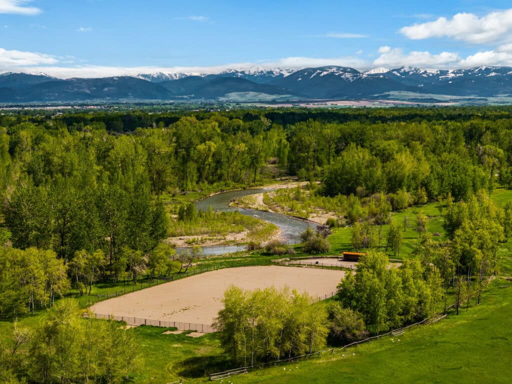 Aerial view of a fenced sandy arena on recreational land, surrounded by lush green trees, a winding river, open grassland, and distant snow-capped mountains under a blue sky with scattered clouds.