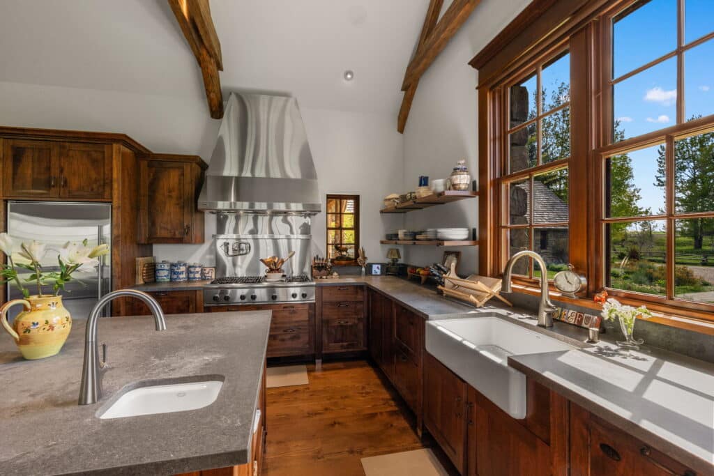 A rustic kitchen with wooden cabinets, gray countertops, a farmhouse sink beneath large windows, stainless steel appliances, and exposed wooden beams. The bright room overlooks green recreational land, perfect for a cattle ranch or land for sale.