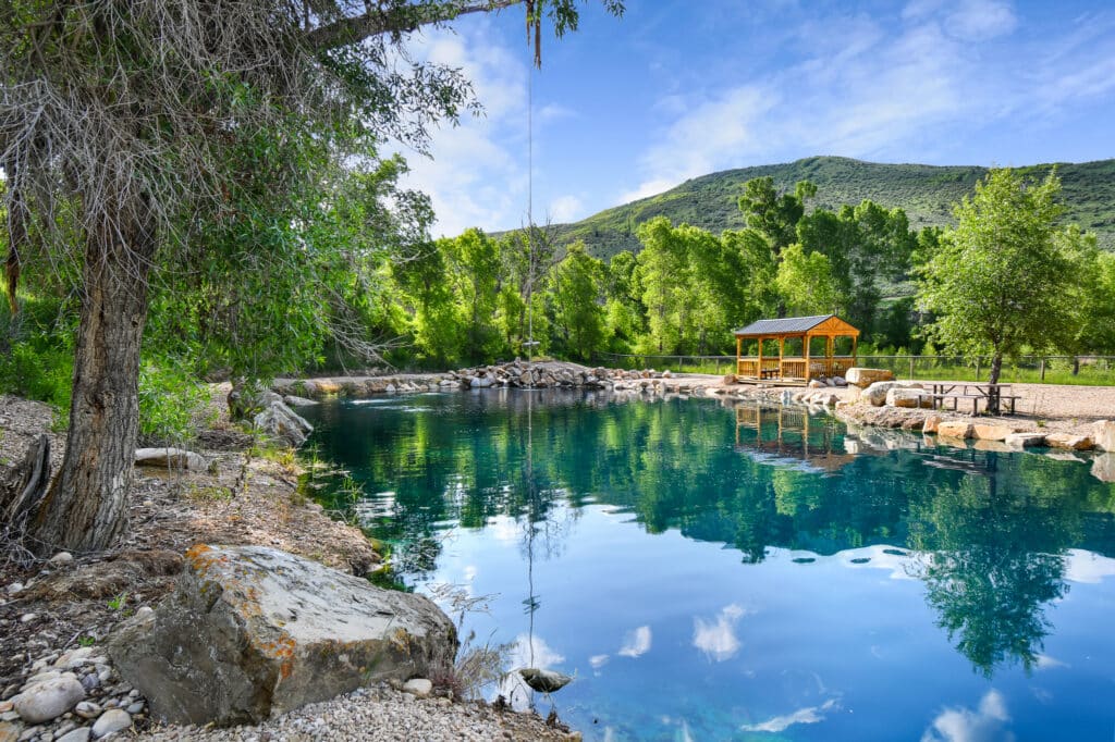A clear blue pond surrounded by trees, rocks, and a gravel shore, with a small wooden shelter and picnic tables in the background—perfect recreational land set against green hills under a partly cloudy sky.