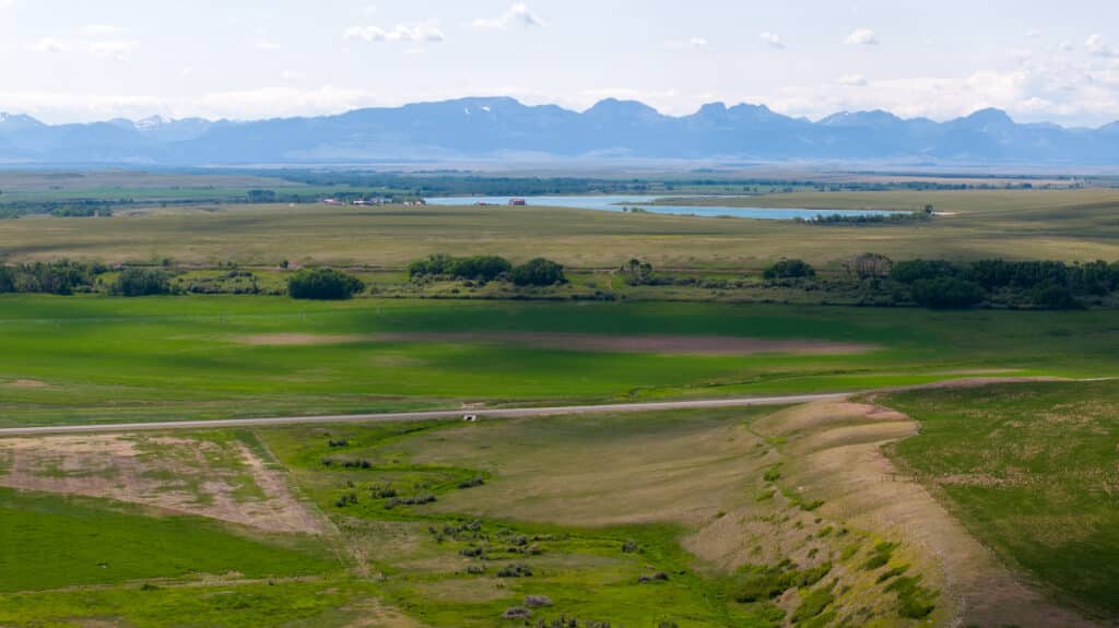 A wide rural landscape with green fields, a winding road, a small vehicle, clusters of trees, a distant lake, and snow-capped mountains under a partly cloudy sky—perfect for a cattle ranch or hunting property.