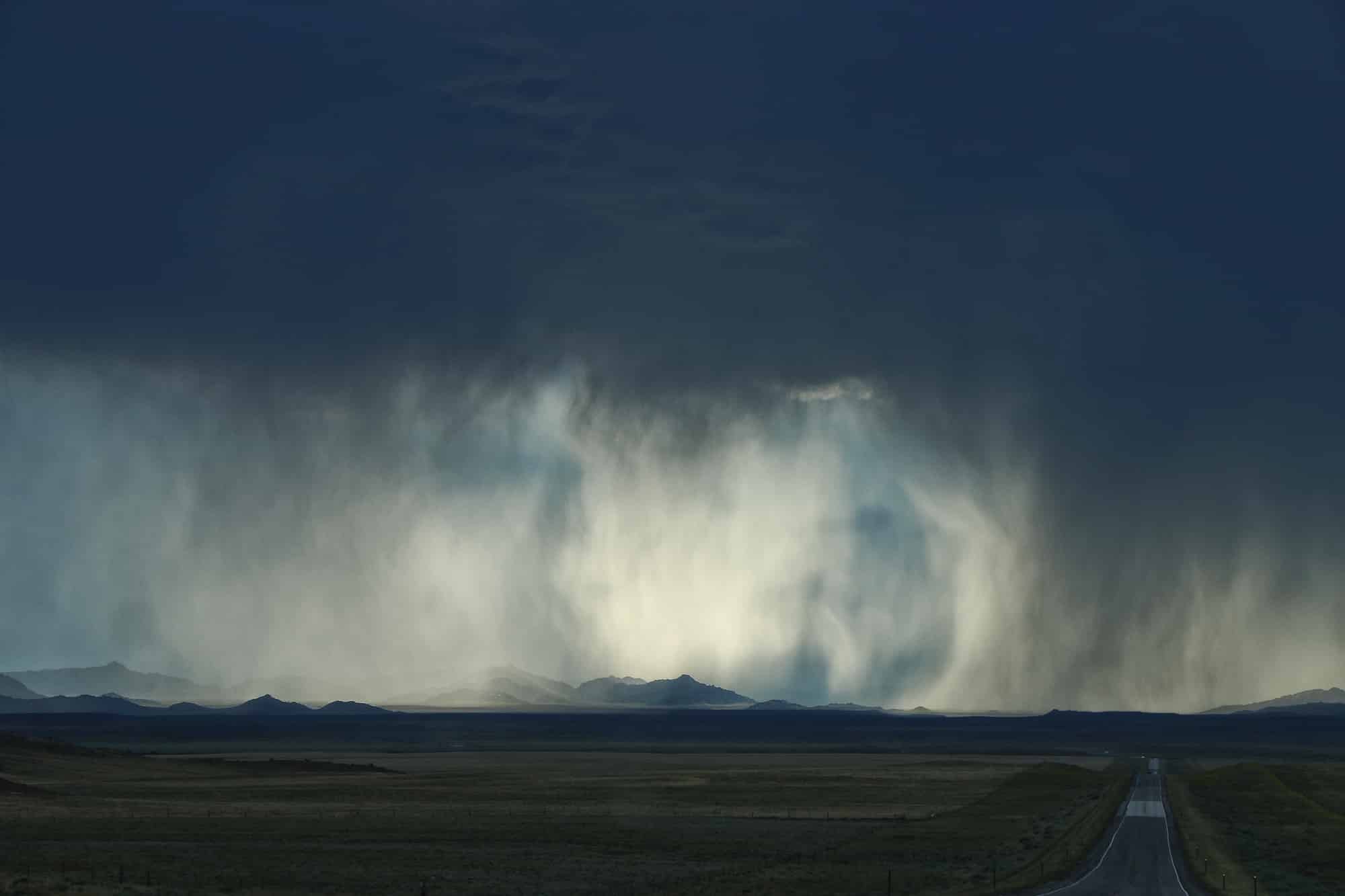 A dark, dramatic sky pours heavy rain over distant mountains as a straight road stretches through open plains toward the storm, highlighting the vast potential of this ranch for sale.