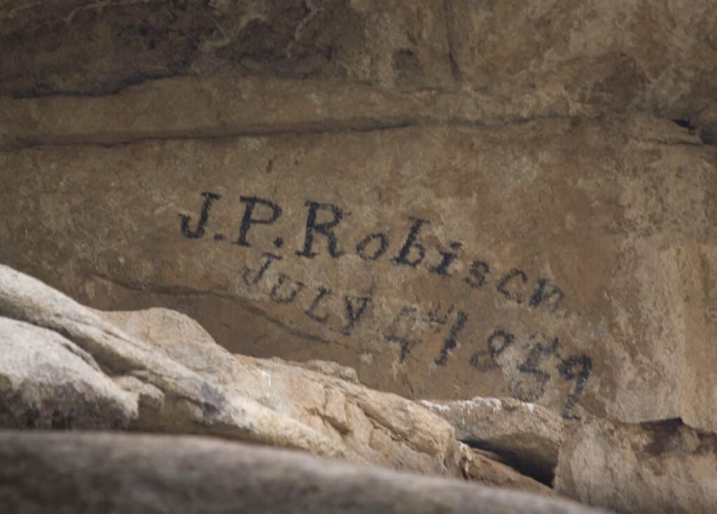 Text “J.P. Robison July 4, 1859” is written in black on a rough, uneven rock surface. The weathered lettering fits seamlessly into the natural stone formations often found on recreational land or a scenic hunting property.