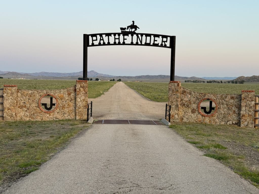 A ranch for sale with a stone wall and metal gate labeled Pathfinder, featuring a horseback rider silhouette. A long dirt road leads through open fields toward distant hills, ideal as a hunting property or cattle ranch.
