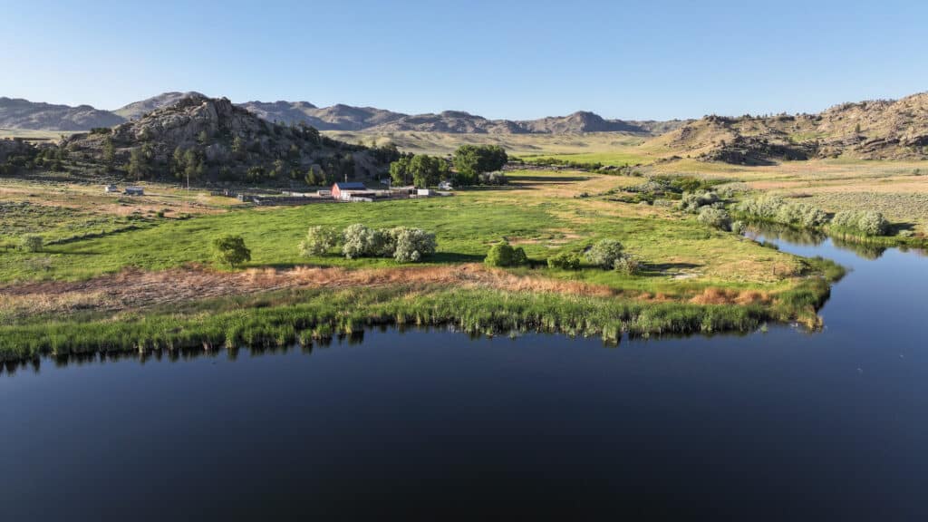A peaceful rural landscape with a river in the foreground, green fields, trees, a few farm buildings, and distant rocky hills under a clear blue sky—ideal for a cattle ranch or those seeking land for sale.