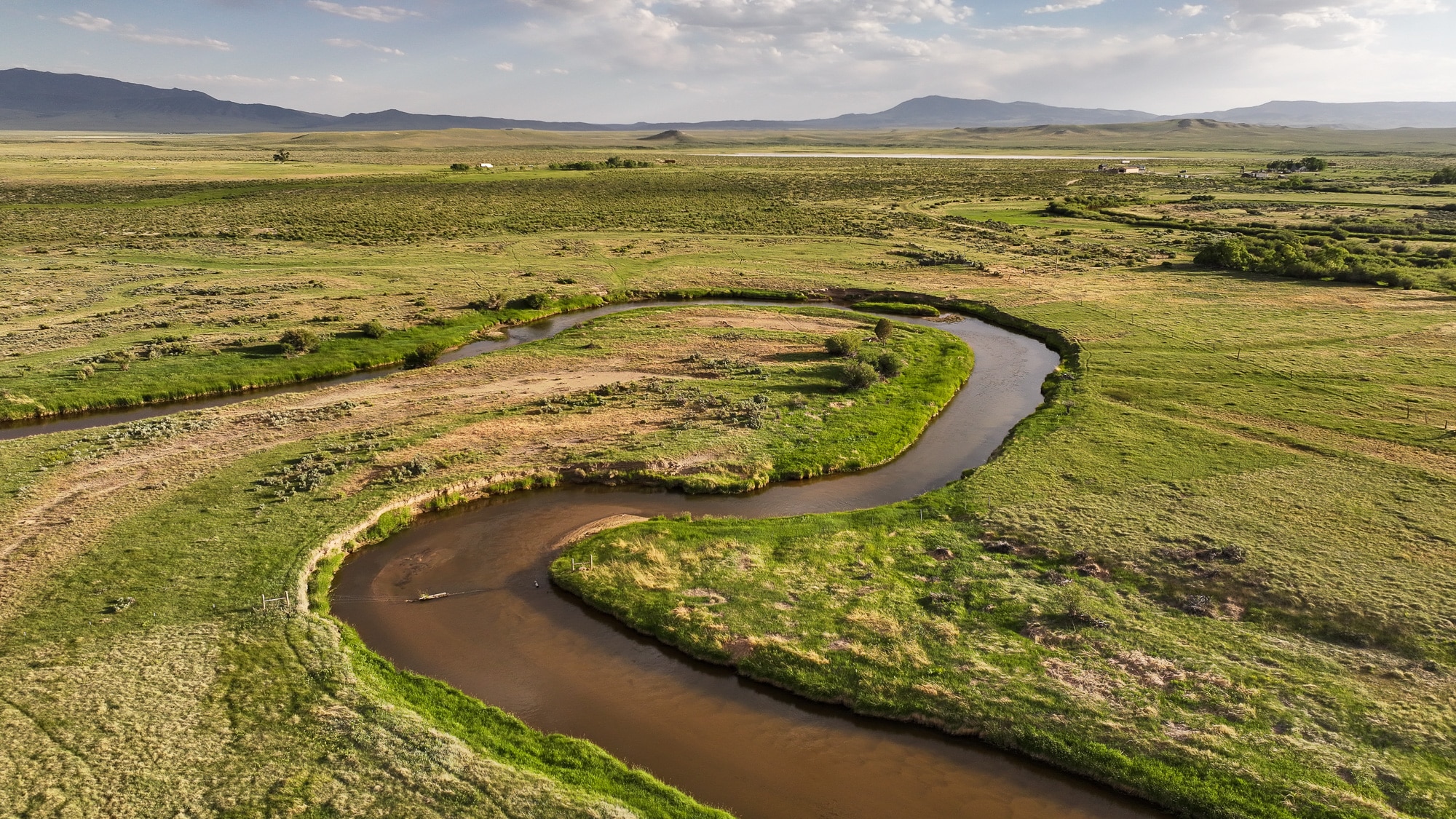 A winding river curves through a vast green landscape with fields and sparse vegetation, set against distant mountains under a partly cloudy sky—an ideal hunting property or ranch for sale.