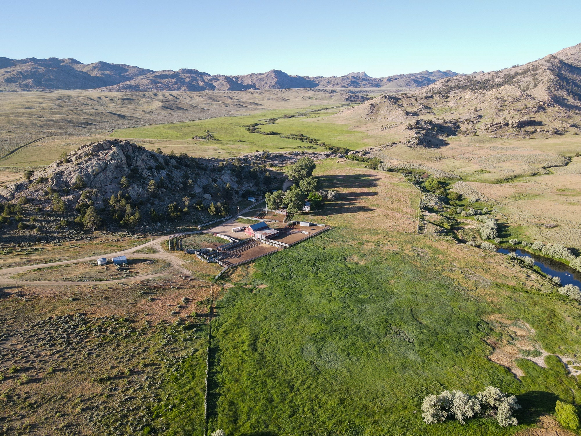 Aerial view of a ranch for sale with barns and fenced areas, surrounded by green fields, rocky hills, and a winding stream, under a clear blue sky.