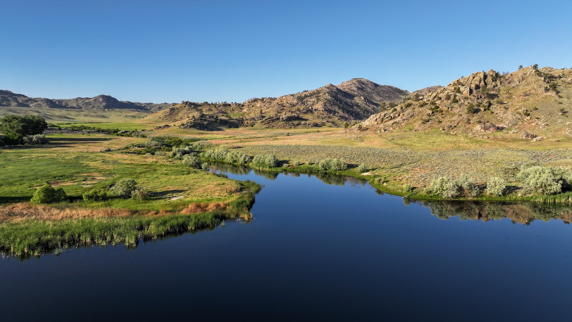 A calm river winds through green fields and grassy hills, with rocky mountains in the background under a clear blue sky—perfect recreational land or hunting property.