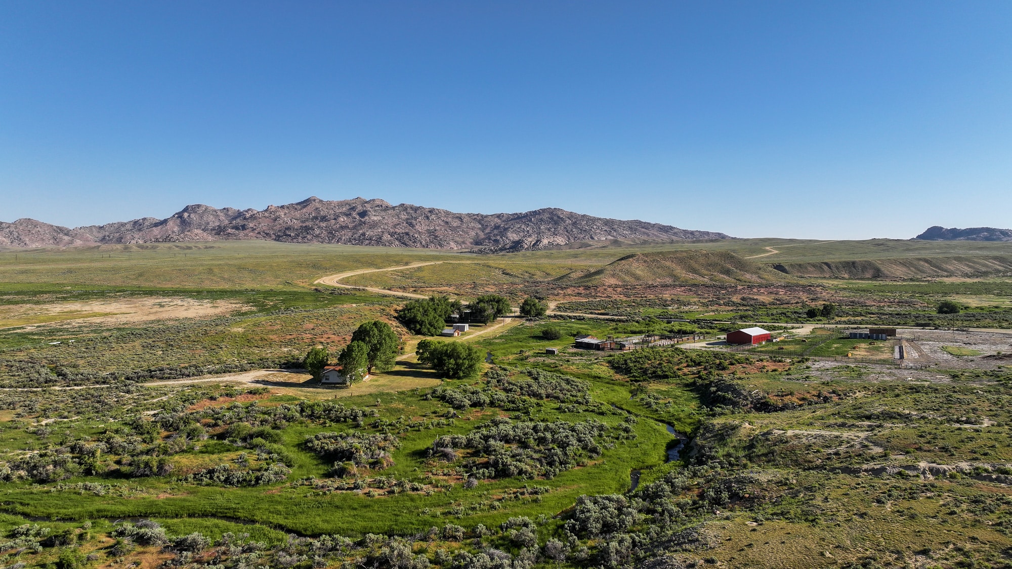 A rural landscape featuring green fields, scattered farm buildings, and a winding dirt road leads to distant rocky hills under a clear blue sky—ideal ranch for sale or recreational land enthusiasts.