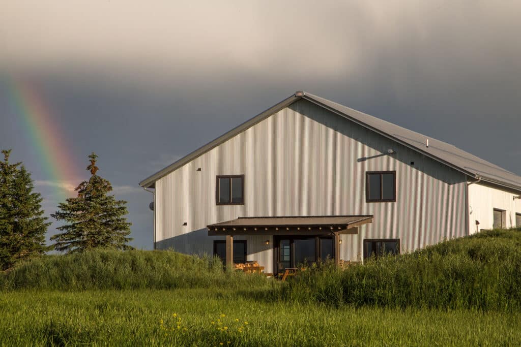 A modern light-gray house sits in a field of tall green grass under a cloudy sky, with a faint rainbow near evergreen trees—an ideal hunting property or ranch for sale.