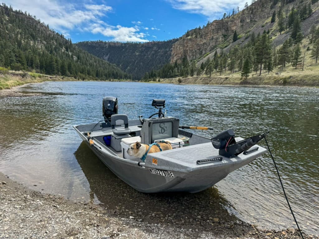 A small gray fishing boat with supplies and a dog aboard is docked on a rocky riverbank, surrounded by forested hills—perfect recreational land or hunting property under a blue sky with scattered clouds.