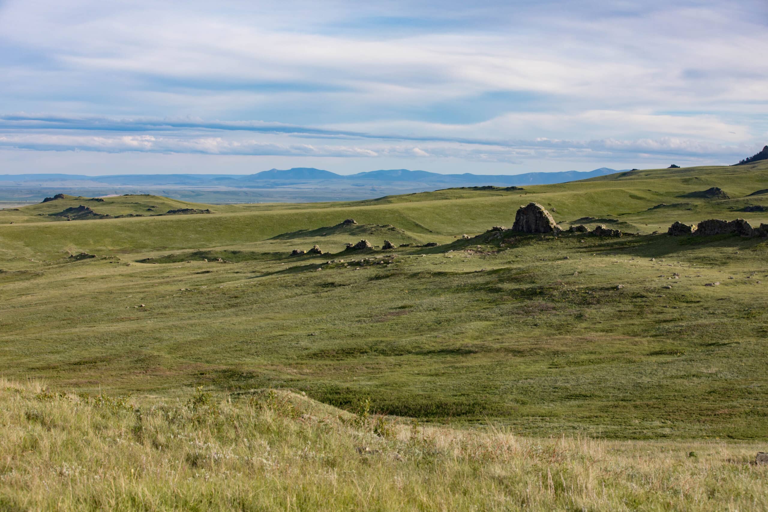 Rolling green grasslands stretch into the distance under a partly cloudy sky, with scattered rocks and distant hills visible on the horizon—ideal as recreational land or potential hunting property.