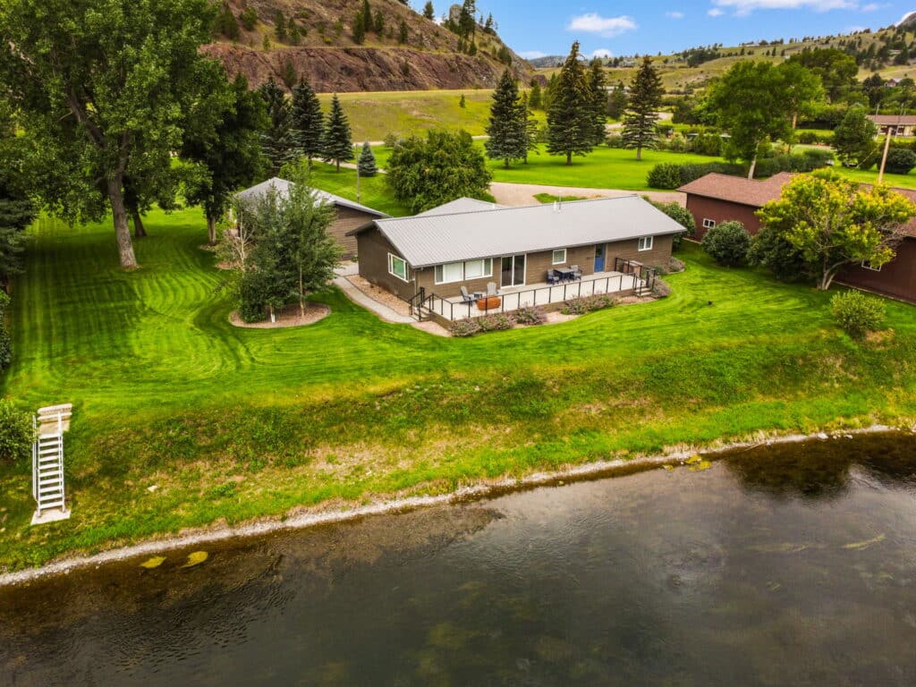 A single-story house with a large deck overlooks a calm river on scenic land for sale, surrounded by lush green grass, trees, and hills in the background under a blue sky.