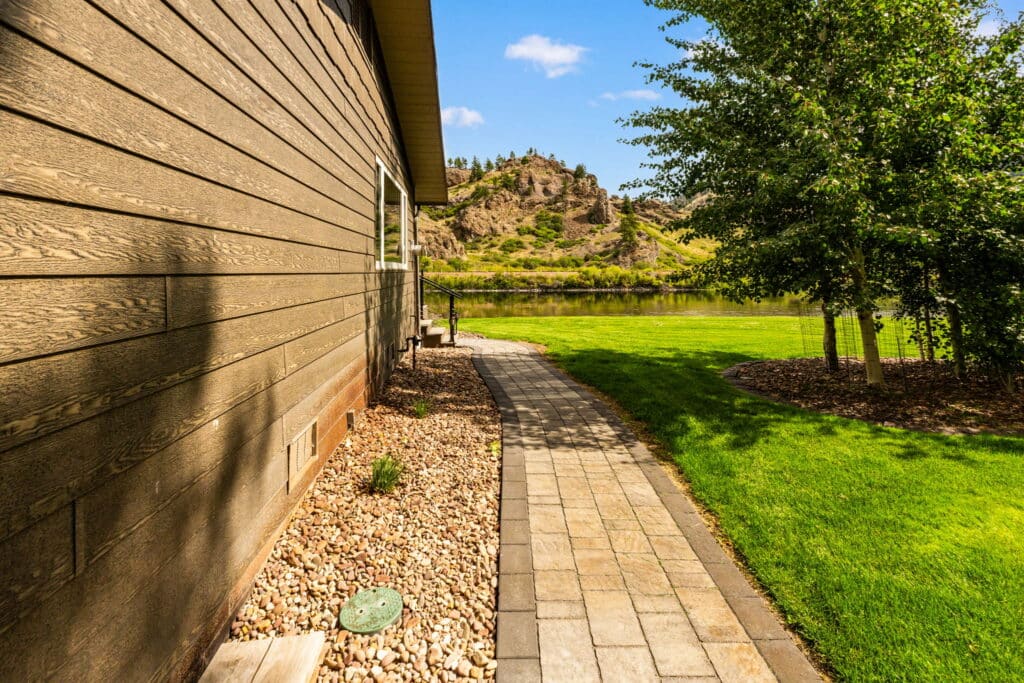 A stone pathway runs alongside a brown house, bordered by rocks and green grass, leading to a scenic lake—perfect for those seeking ranch for sale or hunting property with a rocky hill and trees in the background under a blue sky.