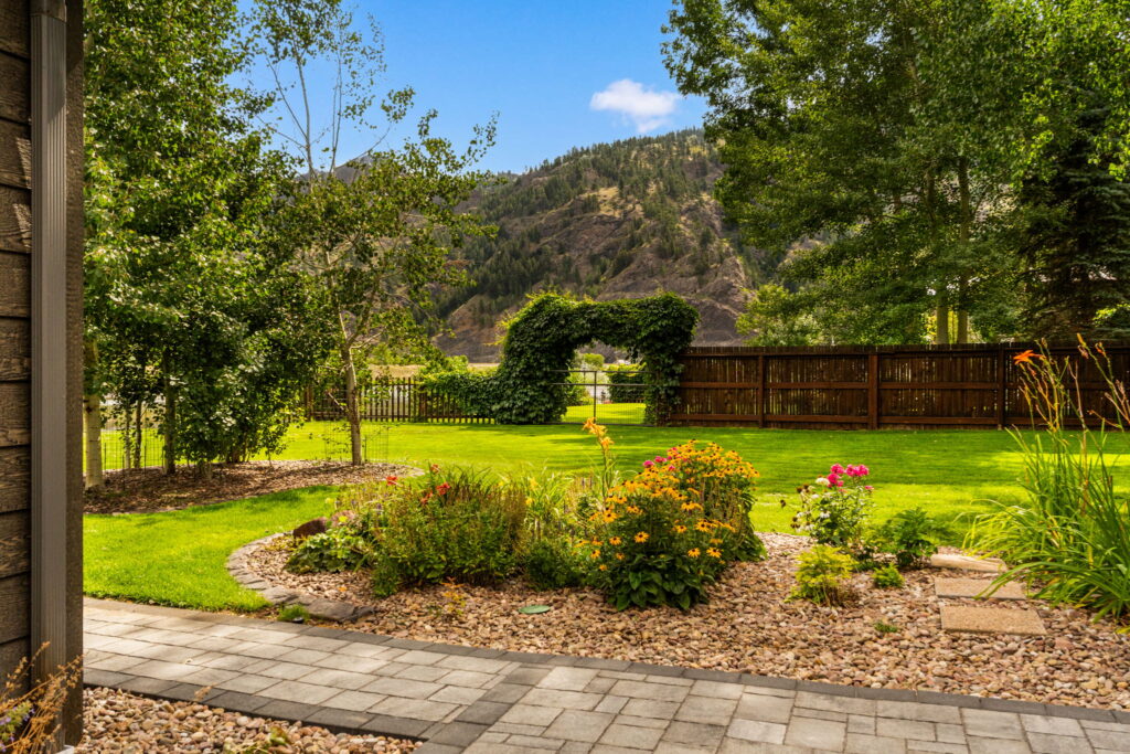 Sunny backyard garden with green grass, blooming flowers, a stone path, wooden fence, and a leafy archway. Trees and hills are visible in the background under a clear blue sky, perfect for those seeking land for sale.