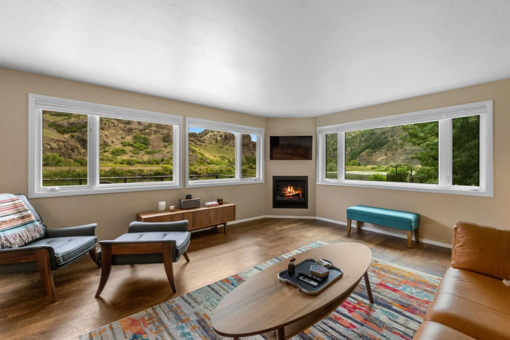 Bright living room with large corner windows showing green hills and a river, featuring a fireplace, modern furniture, a colorful rug, and wood flooring—ideal for those seeking hunting property. A TV is mounted above the fireplace.