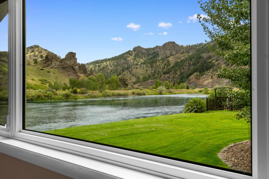 A scenic view through a window shows a calm river, green grass, and rocky hills under a blue sky with a few clouds. Trees line the riverbank, making this ideal recreational land or a picturesque cattle ranch. Part of the window frame is visible in the foreground.