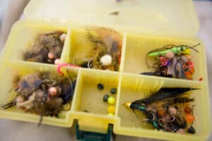 A yellow plastic tackle box with several compartments holds various fishing flies and lures, perfect for preparing for a day out on recreational land or at a cattle ranch. Feathers, fur, hooks, and colorful beads are neatly organized inside.
