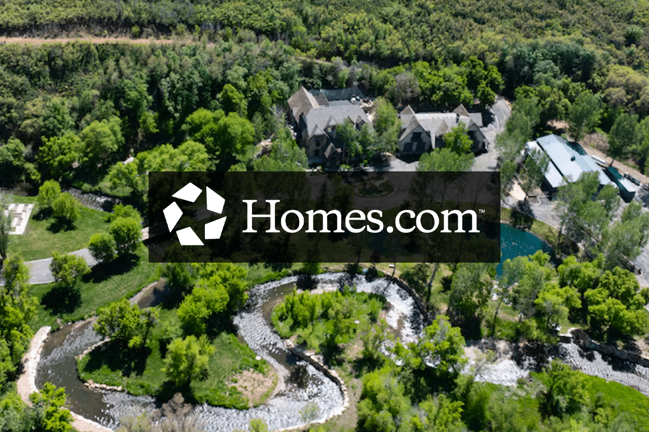 Aerial view of a large, elegant ranch for sale surrounded by dense green trees, a winding river, and landscaped grounds, with the Homes.com logo overlaid in the center.