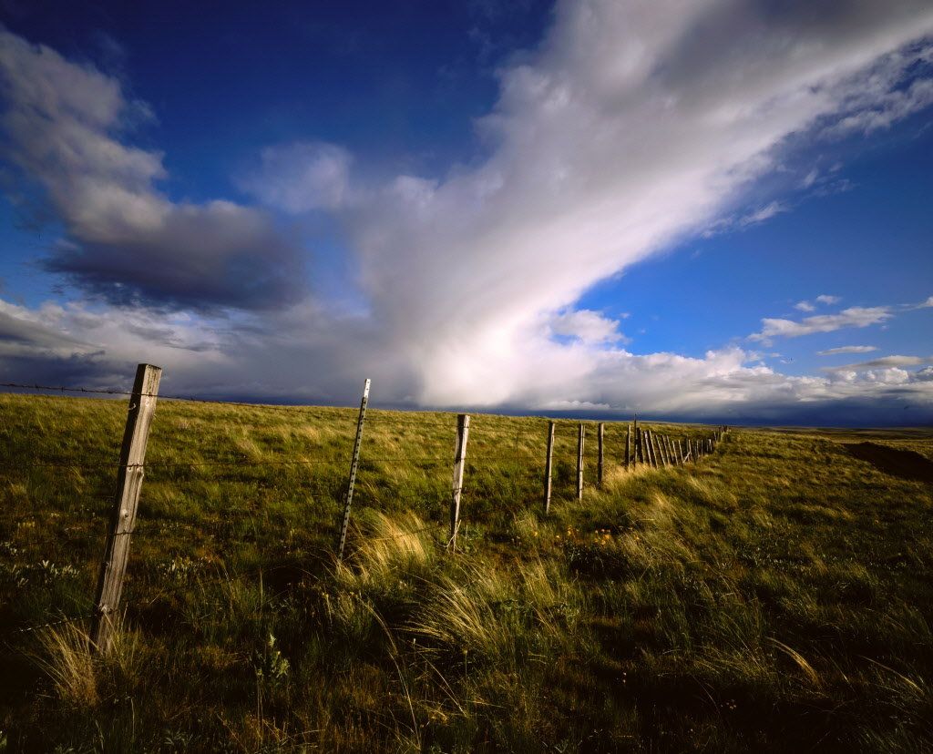 A wooden fence runs diagonally across a grassy field on this scenic recreational land, under a dramatic sky with billowing clouds and patches of blue. The sun casts shadows over the landscape, highlighting the textures of the grass and fence.