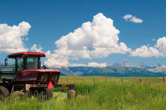A red tractor sits in a grassy field under a blue sky with scattered clouds, with mountains visible in the distance—ideal recreational land or cattle ranch.