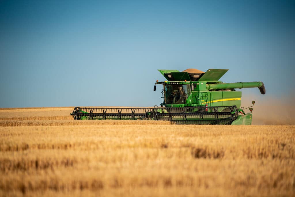 A green combine harvester moves through a golden wheat field under a clear blue sky, harvesting crops on prime land for sale and leaving a trail of dust behind—perfect for those seeking recreational land.