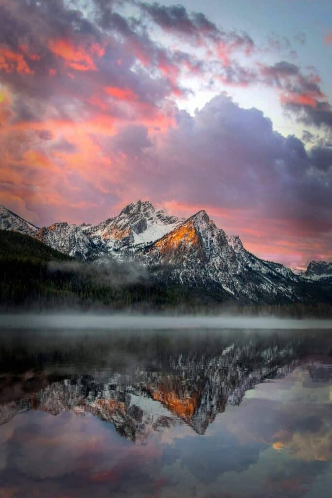 A snow-capped mountain reflected in a calm lake at sunrise, with pink, purple, and orange clouds filling the sky and mist hovering over land for sale perfect for a recreational getaway.