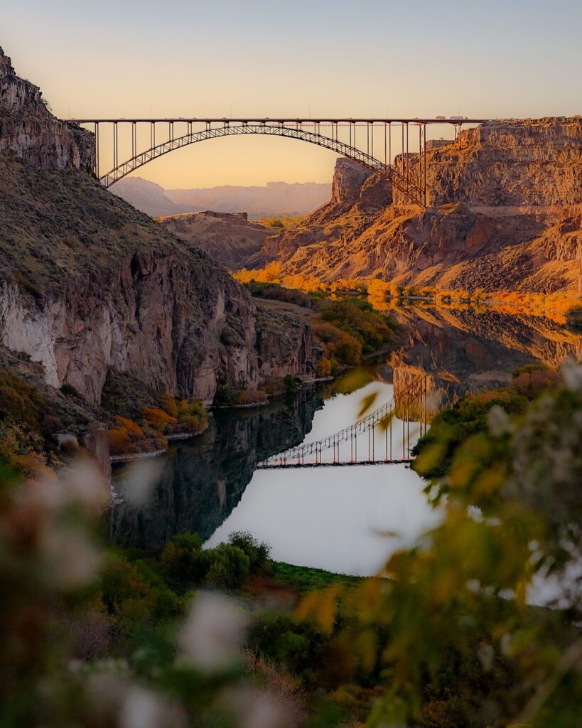 A tall arched bridge spans a deep canyon at sunset, its reflection shimmering on the calm river below, with rocky cliffs and autumn-colored trees. Nearby, hunting property and land for sale stretch along the scenic water’s edge.