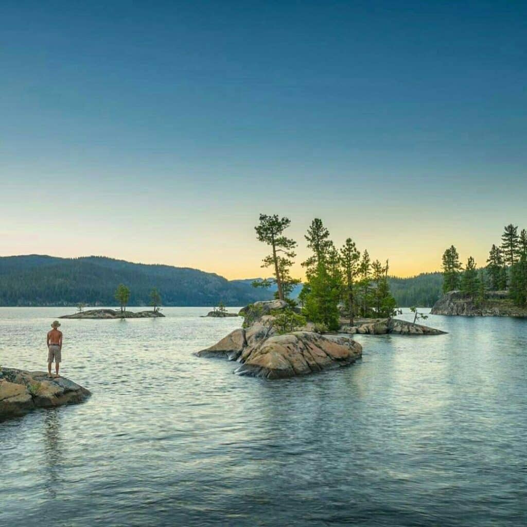 A person stands on a rocky island in a calm lake, surrounded by small, tree-covered islands and distant forested hills under a clear blue sky at sunset—perfect recreational land or hunting property.
