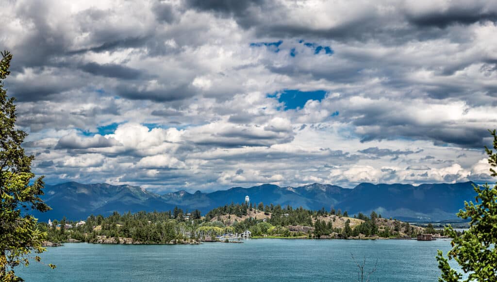 A scenic view of recreational land featuring a lake with a tree-covered island, mountains in the background, and a dramatic sky filled with large, fluffy clouds. Pine trees frame this perfect hunting property on the left and right.