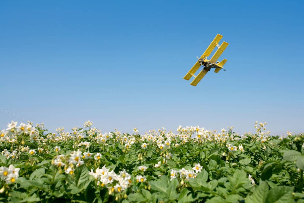 A yellow crop duster airplane flies low over a field of flowering plants, spraying a mist under a clear blue sky—a perfect scene for showcasing land for sale.