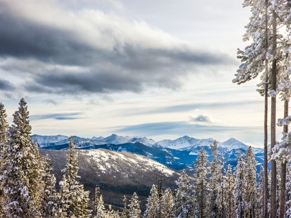 Snow-covered pine trees frame a scenic view of distant, sunlit mountains under a partly cloudy sky. This bright, wintry landscape is perfect for a cattle ranch or recreational land, with rolling hills and dramatic clouds adding depth to the scene.