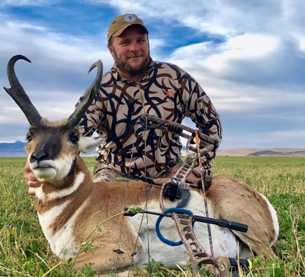 A man in a patterned shirt and tan cap kneels in a grassy field, smiling behind a large antelope he has hunted on prime hunting property, with a compound bow resting on the animal. Mountains and cloudy sky form the backdrop.