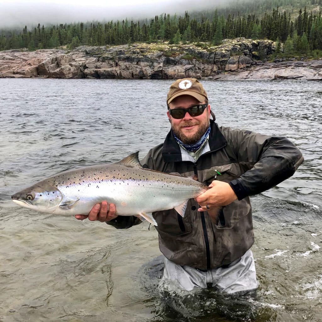 A man stands in shallow water on recreational land, wearing outdoor gear and sunglasses, holding a large fish with both hands. Forested hills and a rocky shoreline appear in the background under a cloudy sky.
