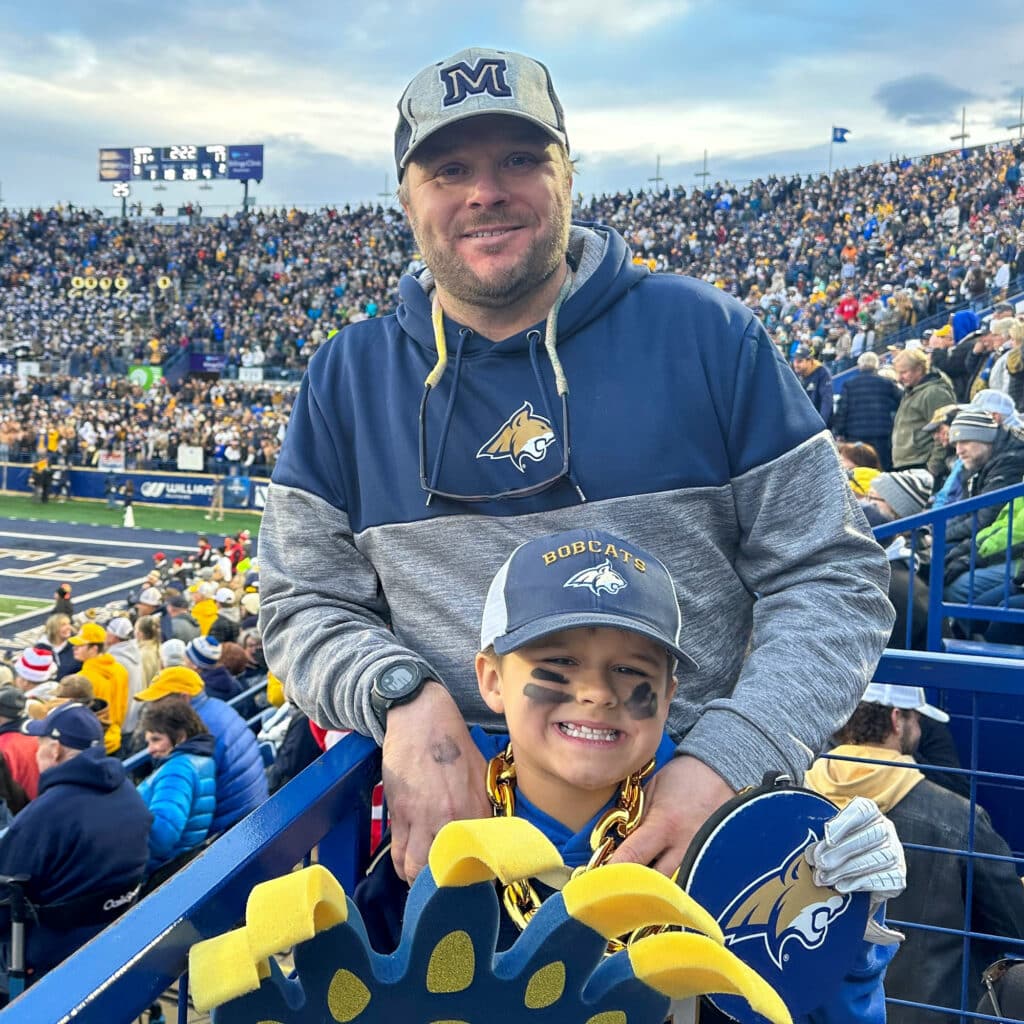 A man and a young boy, both in Montana State Bobcats gear, smile at a crowded football stadium. The boy has face paint, a foam paw, and a gold chain. Fans fill the stands behind them, celebrating after exploring local hunting property.