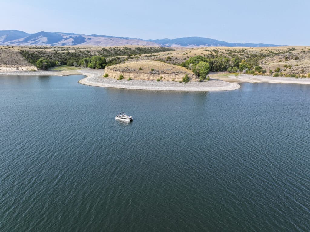 A lone boat floats on a calm lake near a curved shoreline with trees and grassy hills—an ideal setting for recreational land or a tranquil cattle ranch under a clear sky.