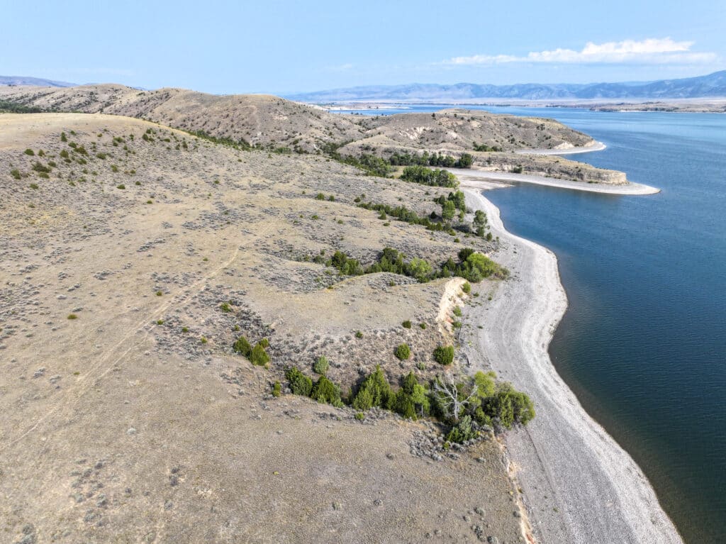 Aerial view of a rocky shoreline with sparse vegetation, gently sloping hills, and calm blue water under a partly cloudy sky. Mountain ranges are visible in the distance, making it ideal as a scenic cattle ranch or hunting property.