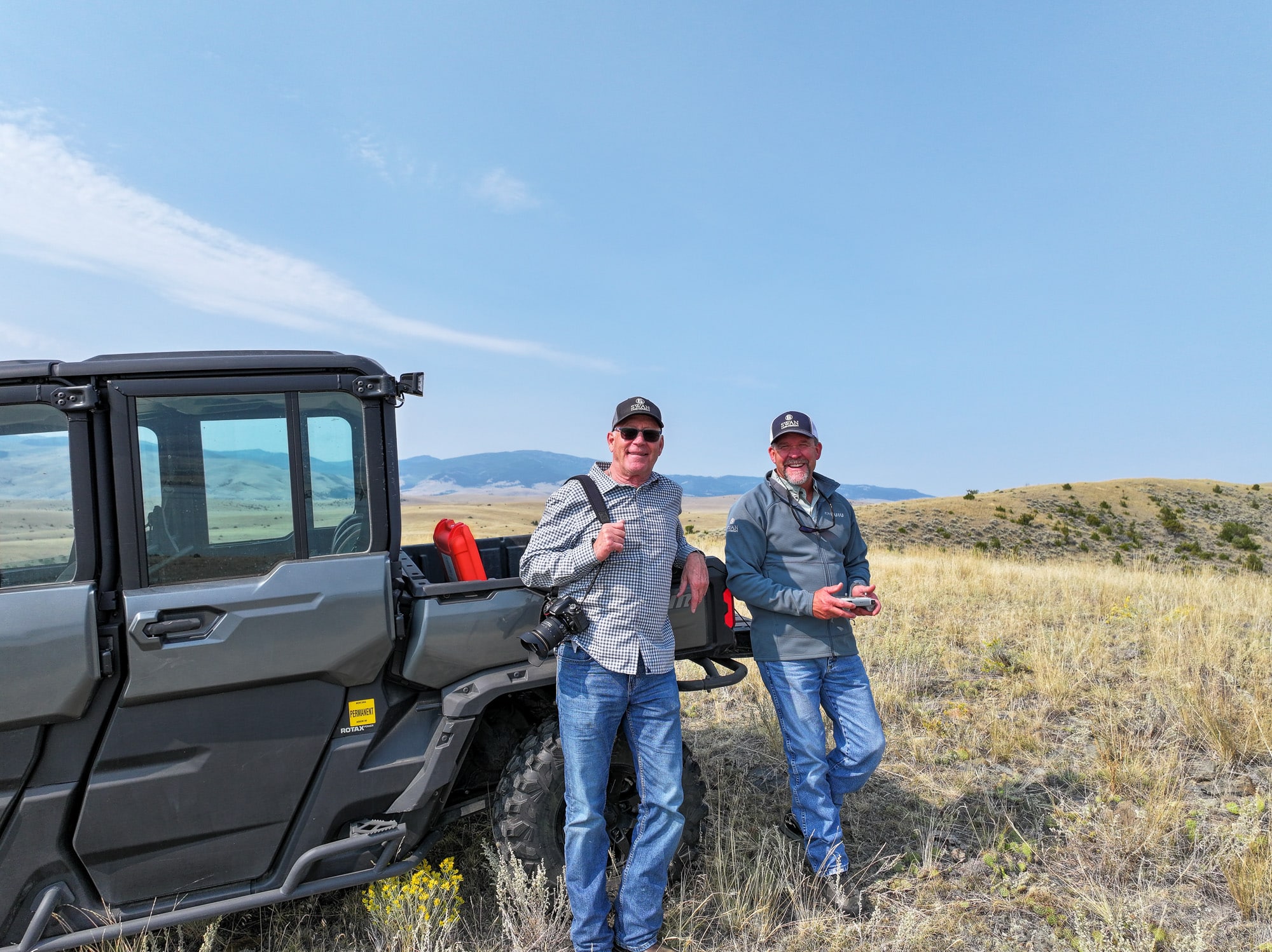 Two men wearing caps and sunglasses stand next to an off-road vehicle in an open grassy field with rolling hills and mountains behind them under a clear sky. One man holds a camera, capturing the beauty of this potential hunting property or cattle ranch.