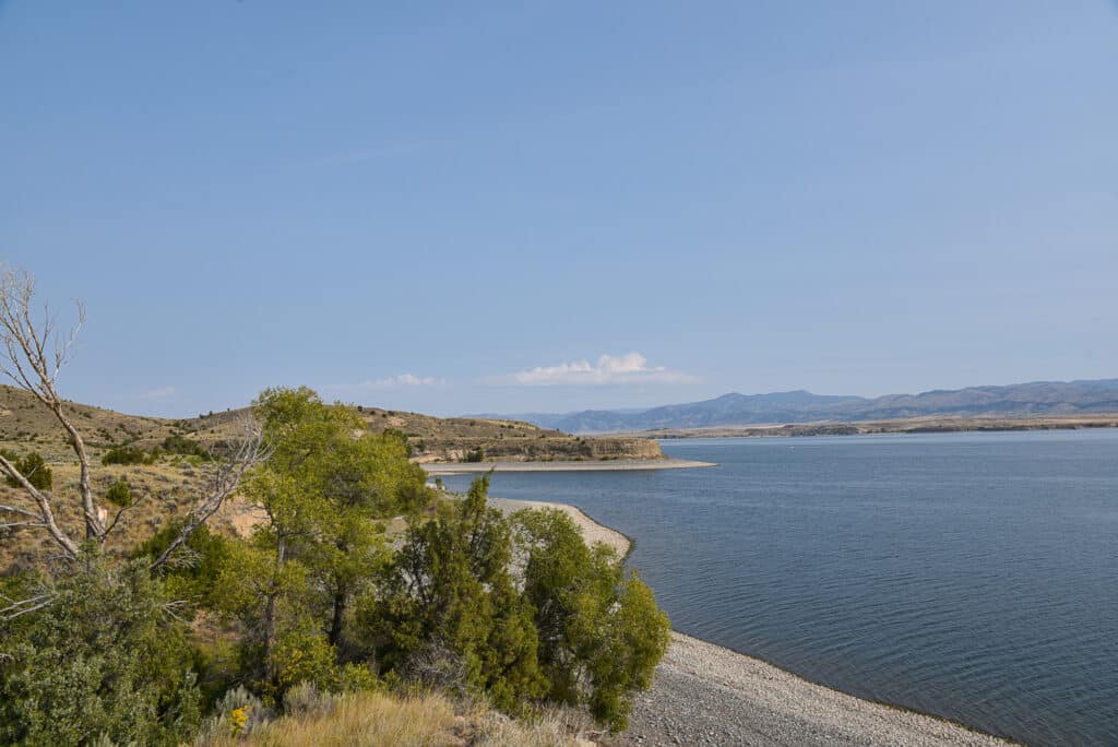 A calm lake curves along a rocky shoreline, bordered by green trees and dry, grassy hills under a clear blue sky with distant mountains—an ideal setting for a hunting property or cattle ranch.