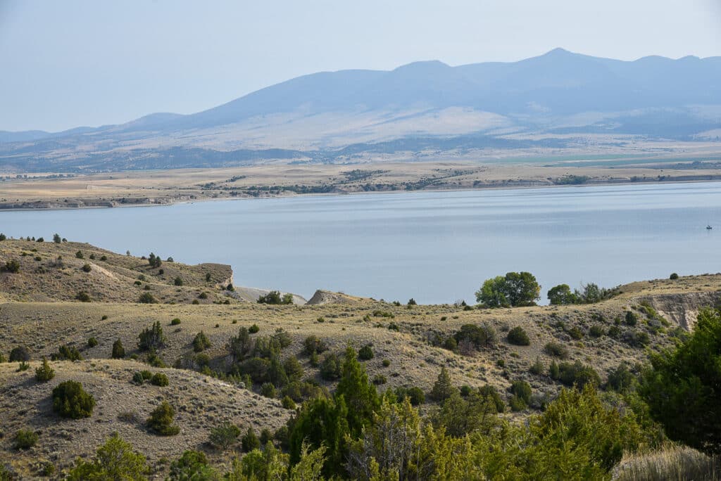 A calm lake surrounded by rolling, grassy hills with scattered shrubs and trees, set against distant mountains under a clear sky—ideal recreational land or cattle ranch opportunities await.