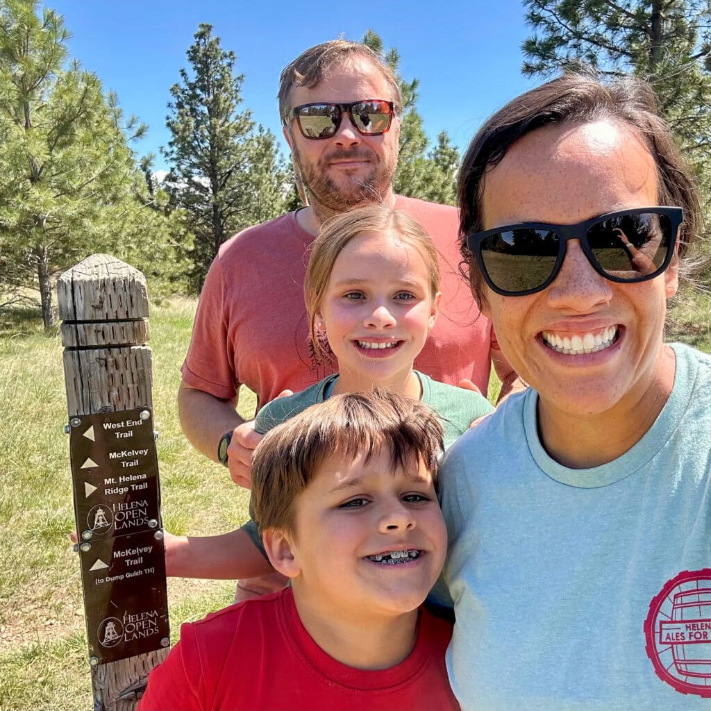 A smiling family of four stands outdoors by a wooden trail sign, surrounded by trees and grass on a sunny day. They wear casual clothes and sunglasses, enjoying a day of hiking near a cattle ranch for sale.