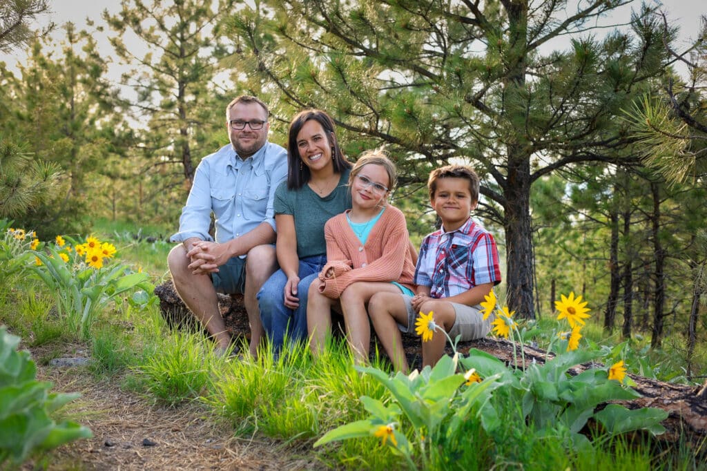A family of four sits on a fallen log in a sunny forest clearing, surrounded by green grass and yellow wildflowers—an ideal spot for your dream hunting property or cattle ranch, with pine trees providing a picturesque backdrop.