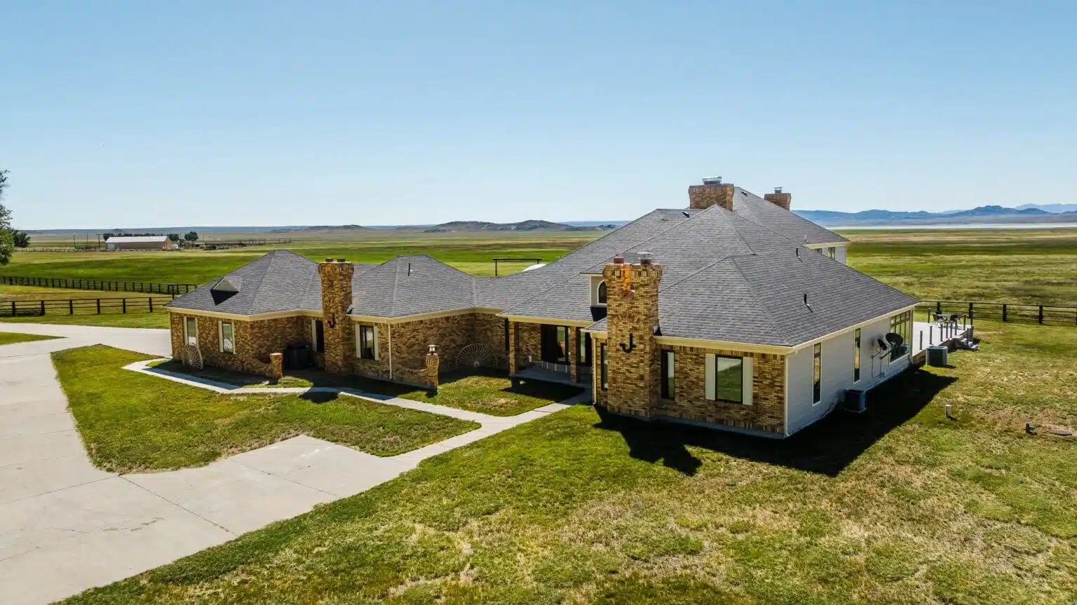A large, single-story brick house with multiple chimneys sits on a spacious grassy lot, ideal as a hunting property, surrounded by open fields under a clear blue sky with distant hills in the background.