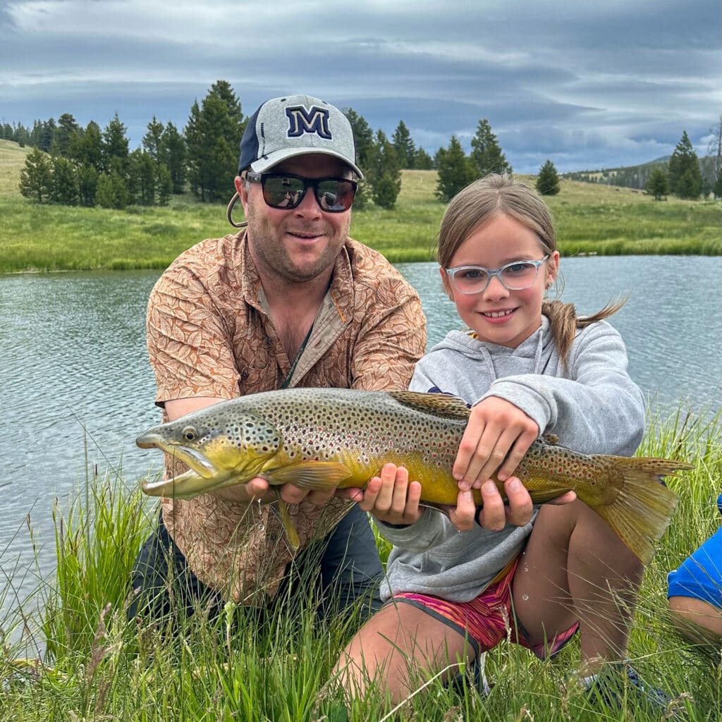 A man and a young girl kneel by a river, both smiling and holding a large brown trout together. Grassy banks, trees, and cloudy skies create an idyllic setting—perfect for enjoying the outdoors on your dream cattle ranch or ranch for sale.