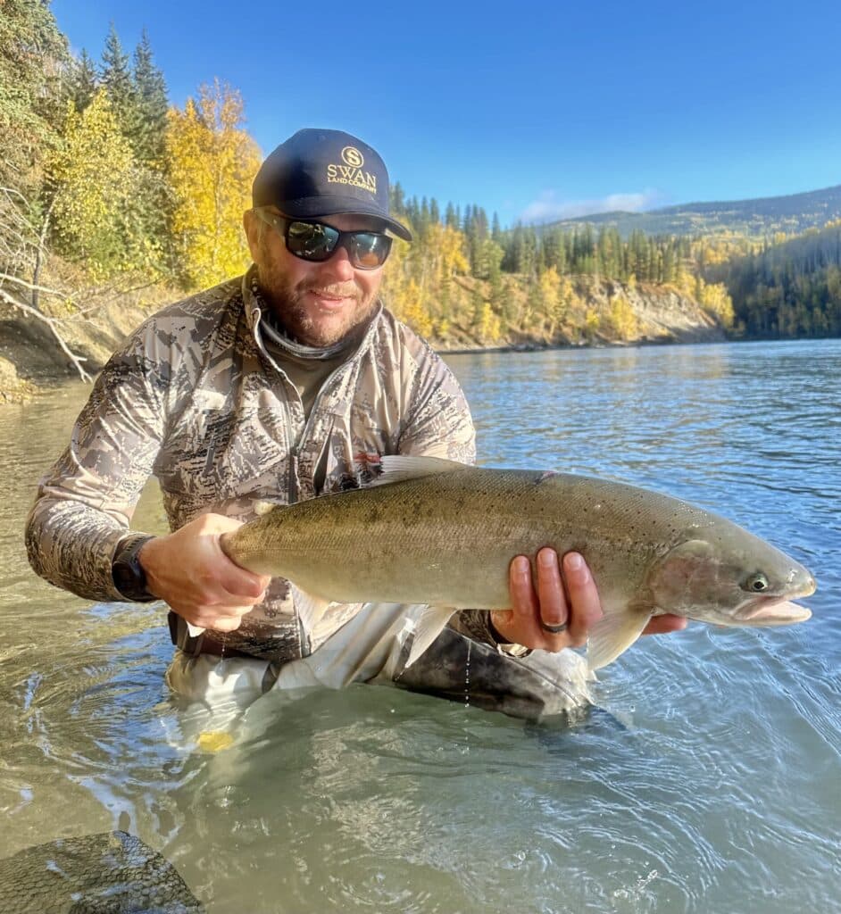 A man in camouflage and sunglasses kneels in a river, smiling with a large fish. Pine trees and autumn foliage line the riverbank under a clear blue sky—perfect scenery for recreational land or hunting property.